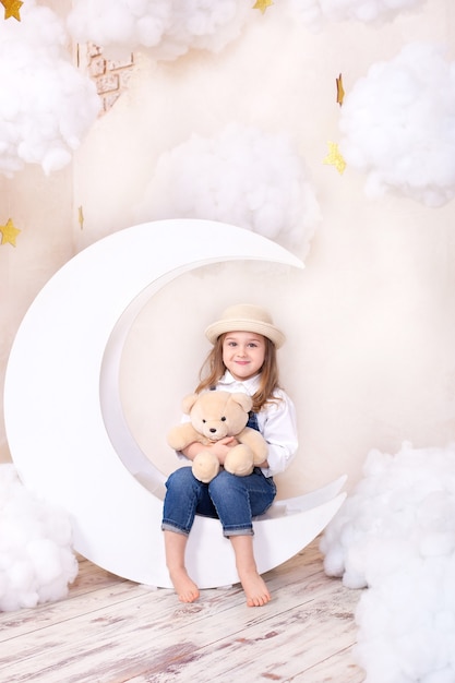 Little girl sitting on moon with clouds with a teddy bear in their hands and playing.