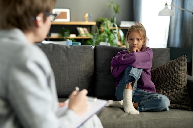 Little girl sitting on sofa in living room and having conversation with psychologist