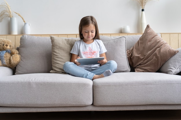 Bambina seduta sul divano a giocare a tavoletta digitale nel soggiorno di casa.