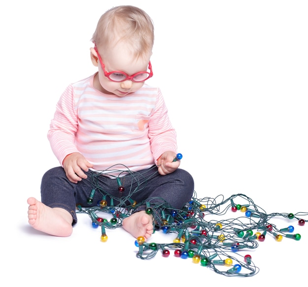 Little girl sitting on white background and playing with Christmas lights. Isolated