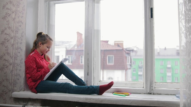 A little girl sitting on the window sill and drawing smiling