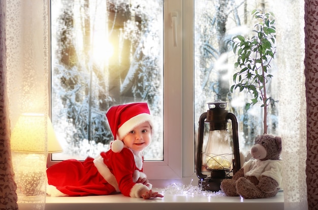 Little girl sitting on a window sill in a suit of Santa Claus