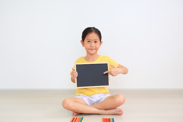 Little girl sitting with holding empty blackboard and pointing on blank board