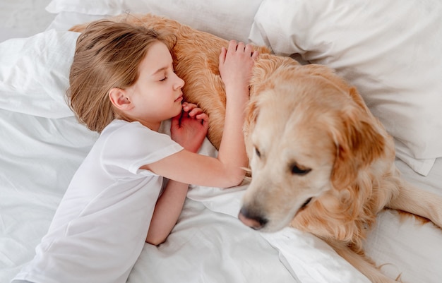 ベッドで寝てゴールデンレトリバー犬を抱き締める少女。朝の時間にペットと昼寝をしている子供。自宅で飼い主と犬