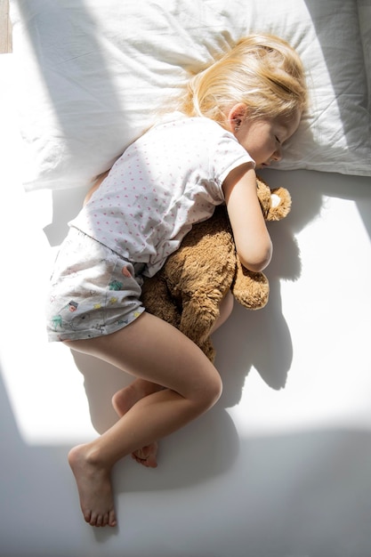 Little girl sleeps hugging a soft toy bear on a white bed Top view flat lay