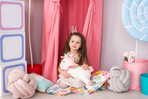 little girl in a smart dress on the background of a beautiful children's room. Toy unicorn in his hands.