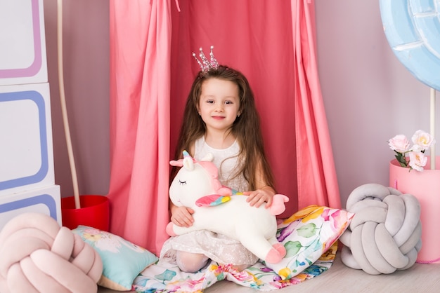 little girl in a smart dress on the background of a beautiful children's room. Toy unicorn in his hands.