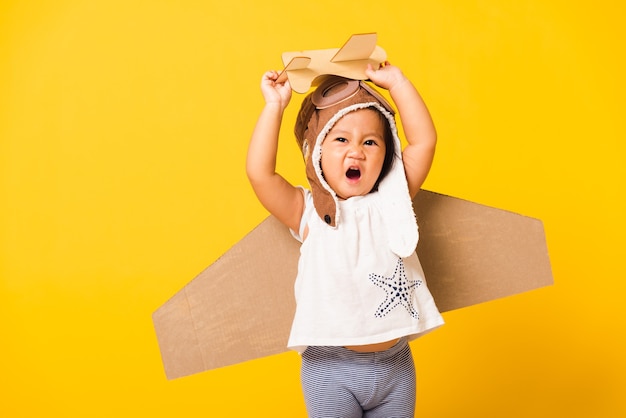 小さな女の子の笑顔はパイロットの帽子の遊びを着て、おもちゃの段ボール飛行機の翼が飛ぶゴーグル