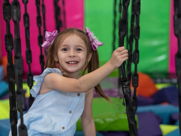 A little girl smiles while sitting in a children's amusement park on a rope wobbling bridge.