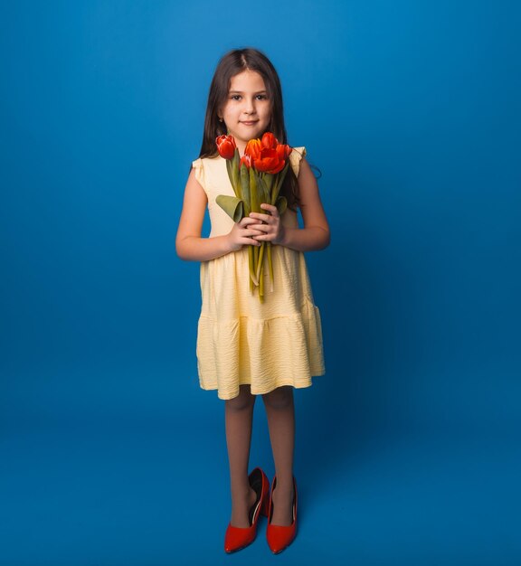 Little girl smiling in a dress and big red shoes holding a bouquet of flowers on a blue background space for text