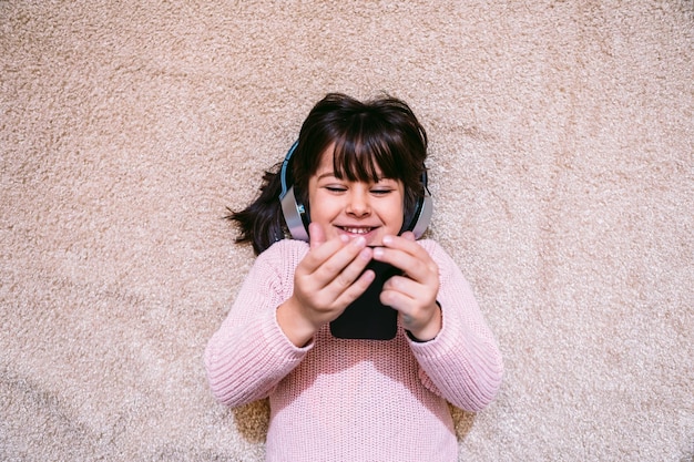 無線ヘッドホンを身に着けている彼女の携帯電話で音楽を聴いたり映画を見たりする敷物の上に横たわっているピンクのセーターで笑っている少女テクノロジーの概念小さな子供と携帯電話