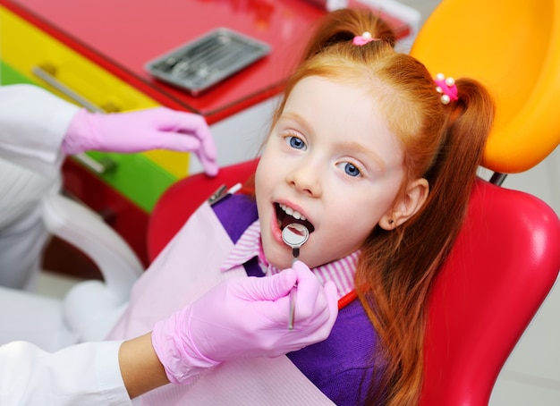 赤い歯科用椅子に笑っている小さな女の子。歯科医は子供の患者の歯を調べます。