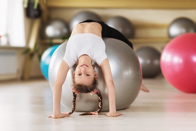 A little girl in sportswear makes a bridge exercise on a fitball in a sports studio