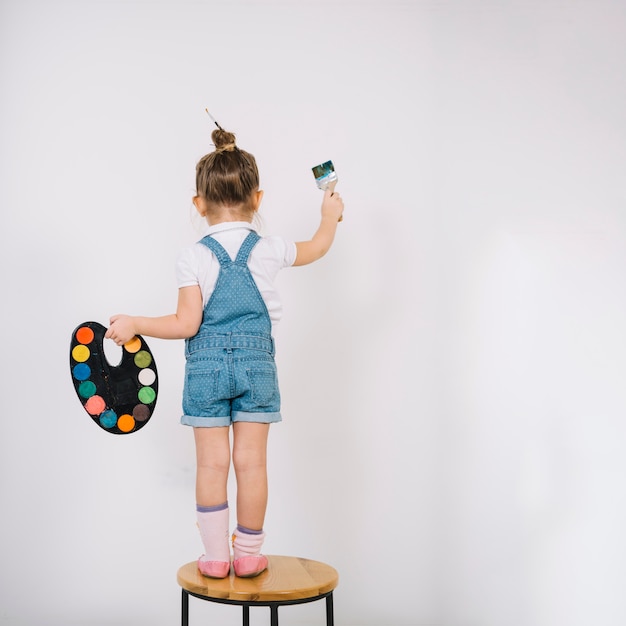 Foto bambina che sta sulla sedia e che dipinge parete bianca con la spazzola