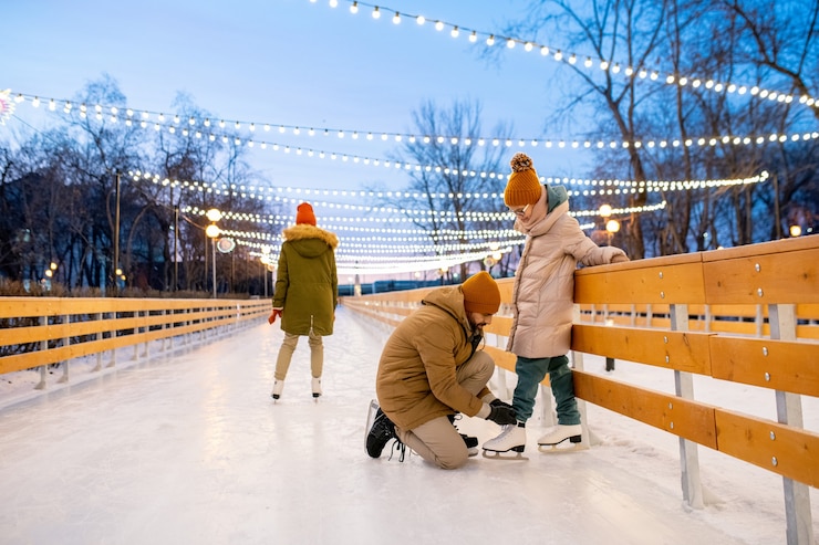 Photo little girl standing on figure skates while her father tying shoelace on her skate during their ride on ice rink