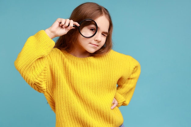 Little girl standing holding magnifying glass and looking at camera with curious expression wearing yellow casual style sweater Indoor studio shot isolated on blue background