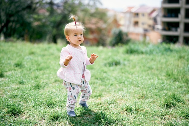 小さな女の子は、建物の表面の木々の間の緑の牧草地に立っています