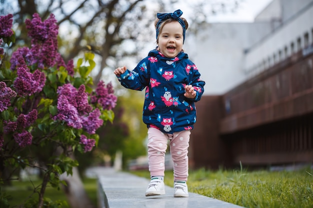 小さな女の子がライラックの緑豊かな茂みの近くに立って、彼女は微笑んで紫色の花を嗅ぎます