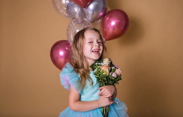 Little girl stands with a bunch of inflatable balloons and holds a bouquet of fresh flowers on a beige background with a place for text