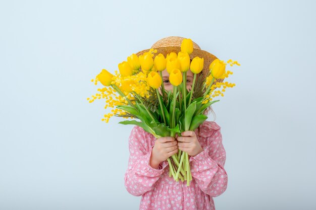 麦わら帽子の少女は、白い背景で隔離の黄色いチューリップの花束を保持します。