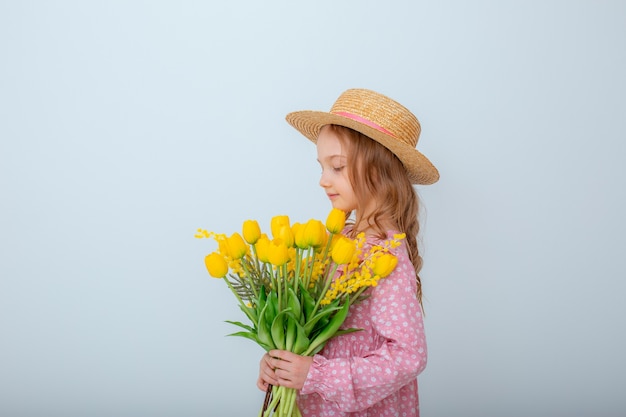 麦わら帽子の少女は、白い背景で隔離の黄色いチューリップの花束を保持します。