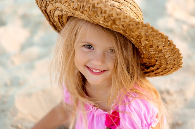 little girl in a straw hat sits on a sandy beach in summer