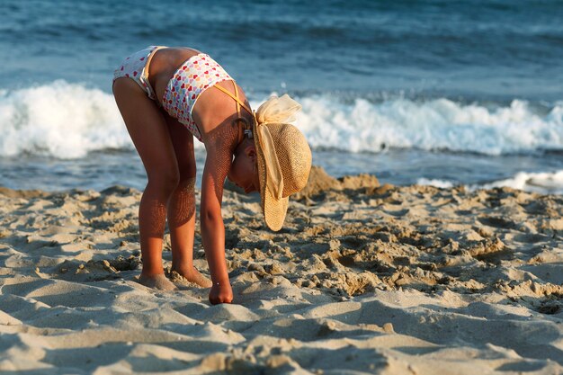 little girl in a straw hat smiles and looks away on a background of the sea