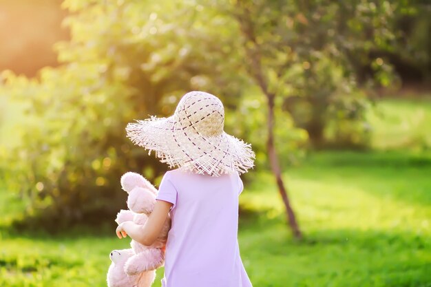 麦わら帽子をかぶった少女は、暖かい黄金の夕日の光に照らされた夏の庭で彼女のおもちゃのクマと一緒に歩き、遊んでいます。