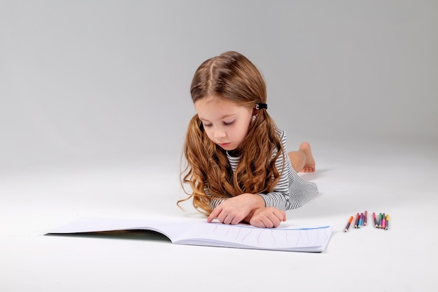 La bambina in un vestito a righe disegna un album sdraiato sullo stile di vita dello sviluppo del bambino del pavimento