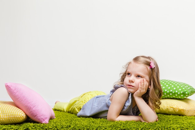 Little girl in a striped t-shirt and yellow jeans lies on a green rug with colorful pillows