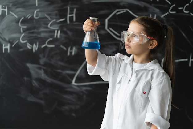 Little girl student with glasses in a laboratory coat stands near the chalkboard holds a chemical fl...