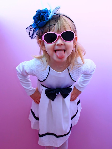 Little girl in stylish hat glasses and fashionable dress showing her tongue Fashionable child expressing her emotions Girl posing in front of camera in modern dress and hat