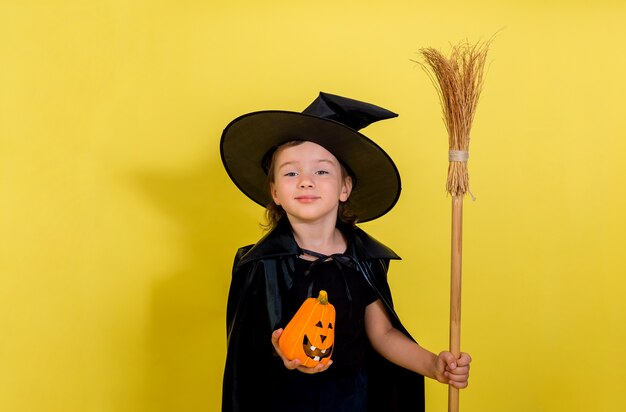 A little girl in a suit witches with a hat with a pumpkin and a broom on a yellow isolated wall with space for text