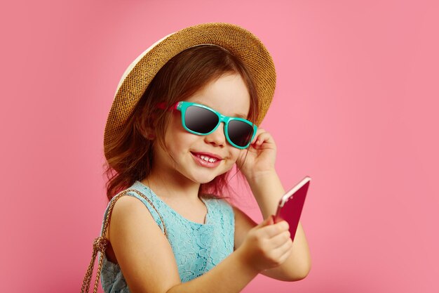 Little girl in summer clothes wearing beach hat sunglasses looking at phone stands on pink isolated