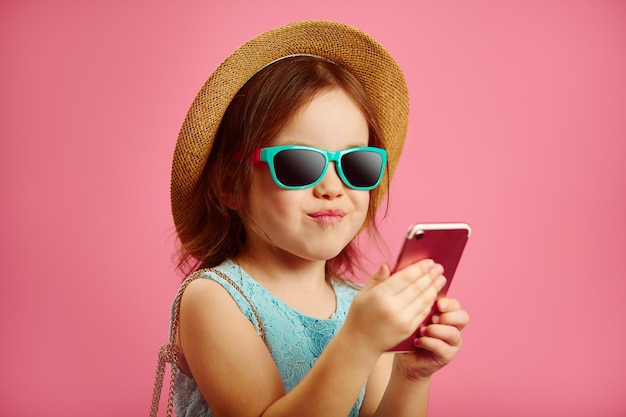 Little girl in summer dress wearing a straw hat and sunglasses looking to phone over pink background