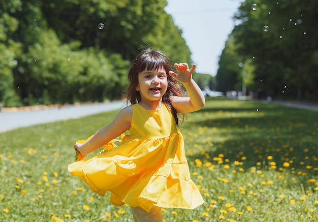 夏の黄色いドレスを着た少女が、タンポポの花輪の公園の芝生でシャボン玉をキャッチします