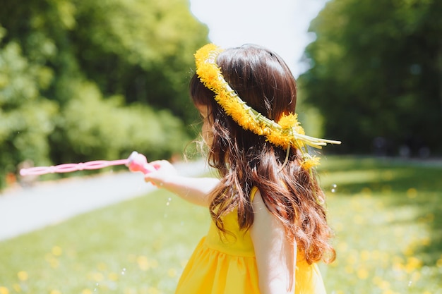 Foto una bambina in un vestito giallo estivo cattura le bolle di sapone sull'erba nella corona di denti di leone del parco