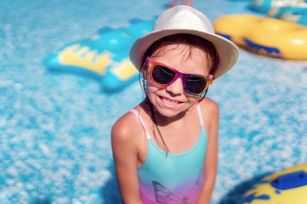 Little girl in Sunglasses and hat with unicorn in outdoor swimming pool of luxury resort on summer vacation on tropical beach island