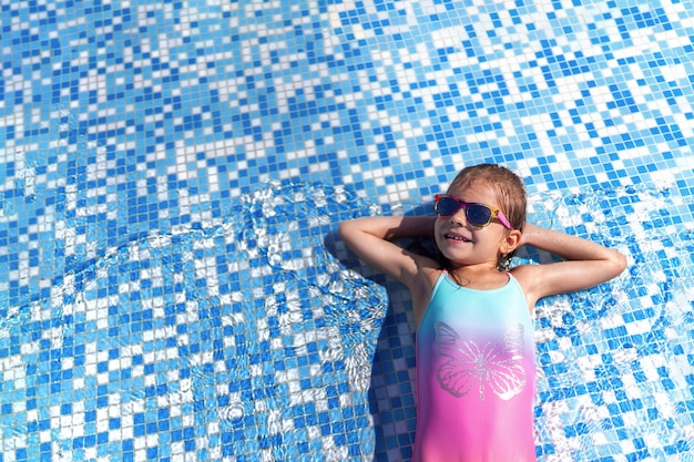 Bambina in occhiali da sole e cappello con unicorno nella piscina all'aperto del resort di lusso in vacanza estiva sull'isola tropicale della spiaggia