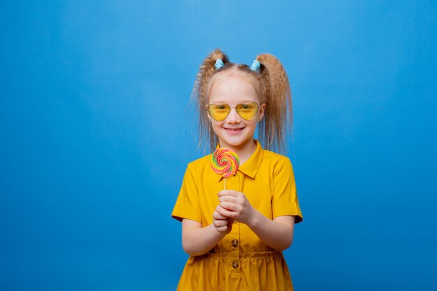 A little girl in sunglasses holds a lollipop on a blue background