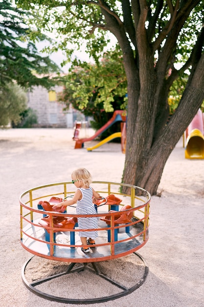 Little girl in sunglasses stands on a turntable swing back view