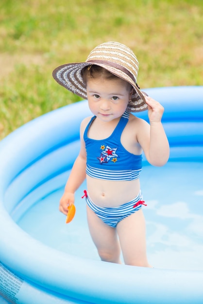 Маленькая девочка в купальнике играет и плавает в надувном бассейне на лужайке в саду