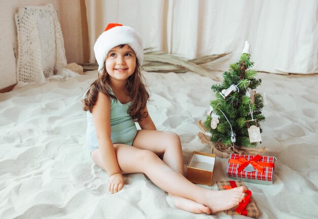 A little girl in a swimsuit and a Santa Claus hat is decorating a Christmas tree and sitting on the sand during the Christmas holidays copy space