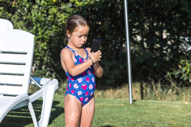 수영장 가장자리에 수영복을 입은 어린 소녀가 휴대전화를 보고 어린 시절 어린이 스마트폰 기술 휴가 인터넷과 재미있는 개념을 보고 있습니다.