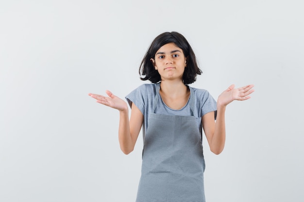 Little girl in t-shirt, apron showing helpless gesture and looking confused , front view.