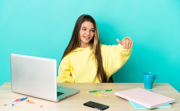 Маленькая девочка в столе с ноутбуком на изолированном синем фоне, показывая большой палец вверх жест