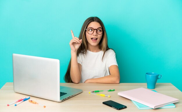 Little girl in a table with a laptop over isolated blue background intending to realizes the solution while lifting a finger up