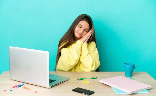 Little girl in a table with a laptop over isolated blue background making sleep gesture in dorable expression