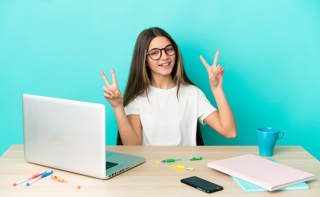 Little girl in a table with a laptop over isolated blue background showing victory sign with both hands