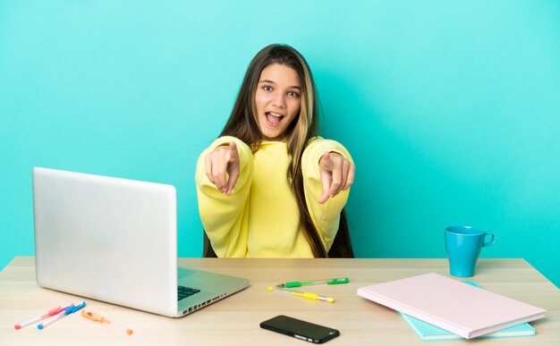 Little girl in a table with a laptop over isolated blue background surprised and pointing front